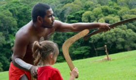Australian Aboriginal warrior man teaching young Australian girl (female age 05) how to throw a boomerang during a cultural show in far tropical north of Queensland, Australia.